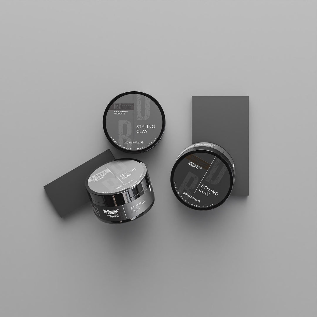 Two jars of hair styling clay and two rectangular clay bars arranged on a smooth light gray surface. The jars have black lids with white text reading 'STYLING CLAY' and show the brand name 'Le Pommier'.